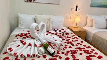 Een hotelkamer met een bed versierd met witte handdoeken en rozenblaadjes, een fles wijn en twee glazen. Twee schilderijen hangen aan de muur, waarvan één een strandafbeelding en de ander een tropisch landschap toont.