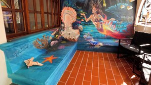 A room with a mural of a mermaid and underwater scene, a window with blinds, a tiled floor, and a chair. - Rachel’s Cove Room 2, Rachel's Cove - Room 2 The Mermaid and The Pearl Hotel (Isla Mujeres)