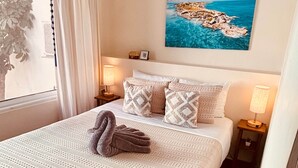 A neatly made bed with a swan pillow, a painting of a coastal scene, and a window with curtains.