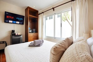 A hotel room with a bed, a flat-screen TV mounted on the wall, a wooden wardrobe, a window with curtains, and a small table with a coffee maker and cups.