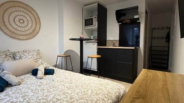 A bedroom with a bed, a wooden table, and a kitchenette in the background. - Le - Studio - Louana (Aubusson)