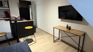 A compact living space with a kitchenette, a sofa, a small dining table, and a TV mounted on the wall. - Le - Studio - Louana (Aubusson)