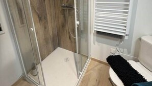 A modern bathroom with a glass-enclosed shower, wooden panel walls, a toilet with a black bath mat, and a white radiator. - Le - Studio - Louana (Aubusson)