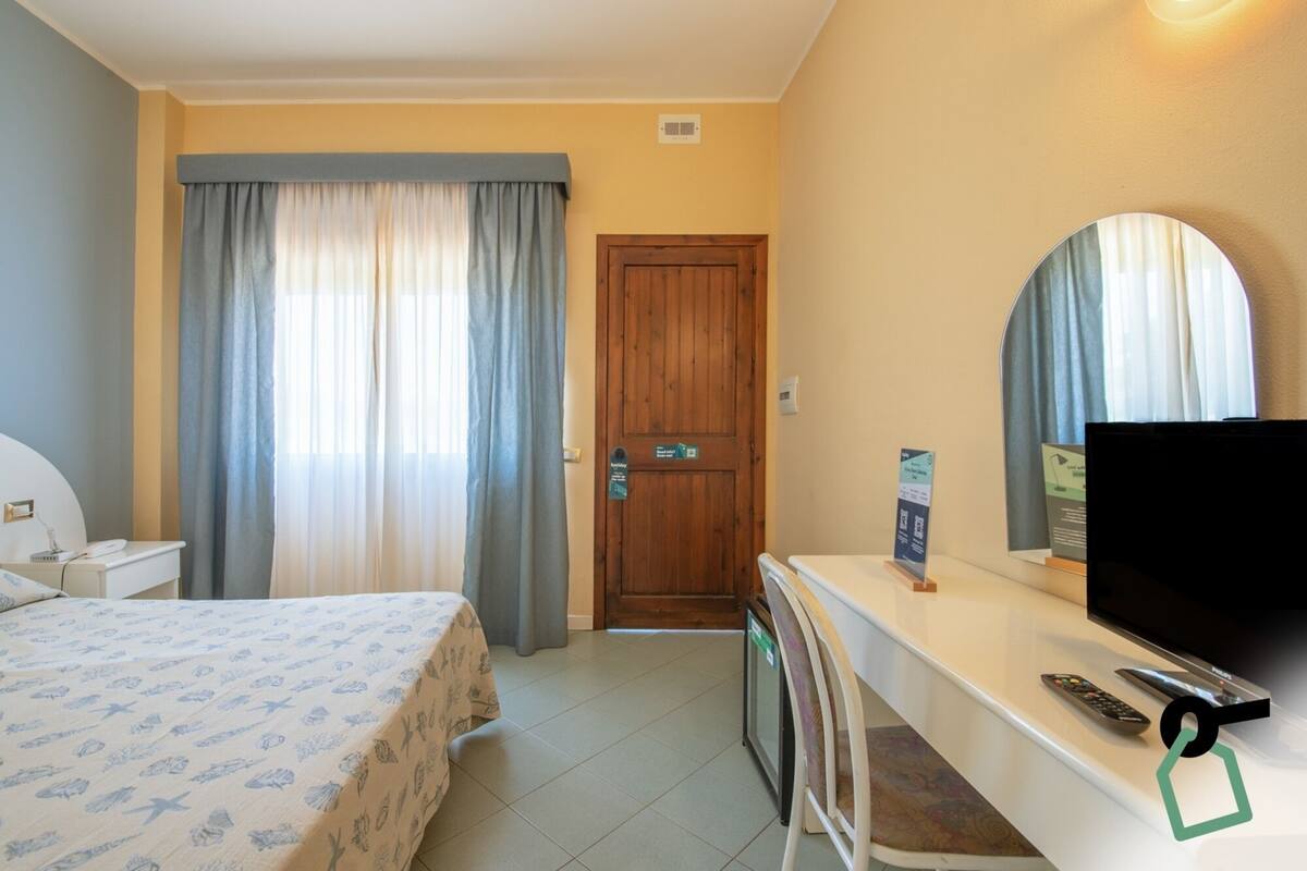 Una habitación de hotel con una cama, un escritorio con televisor, un espejo y una puerta.