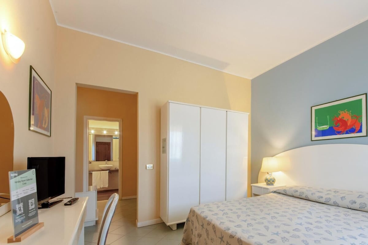 Habitación de hotel con cama, escritorio con computadora, armario y baño visible a través de una puerta abierta.