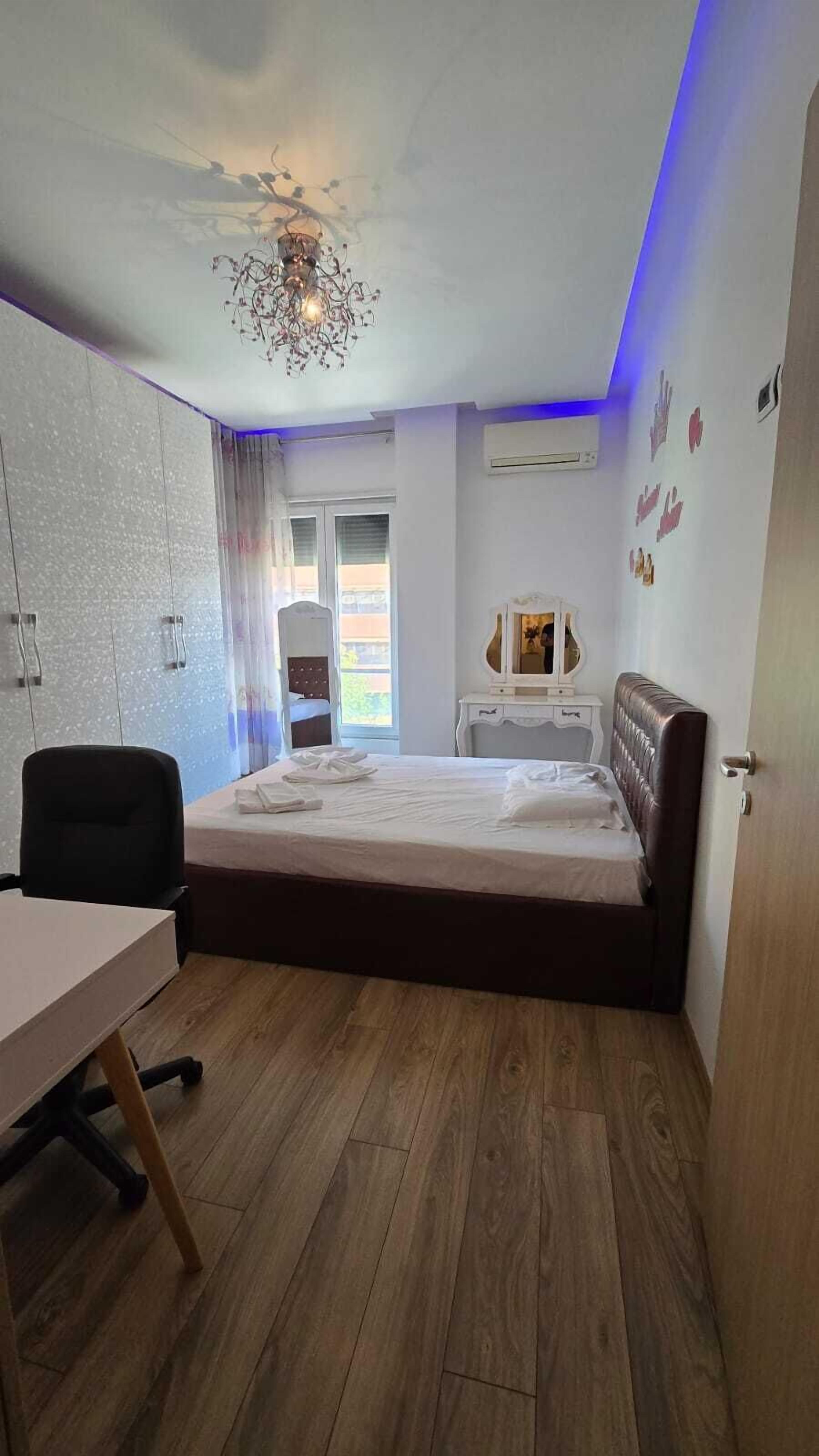 A bedroom with a bed, a desk, and a chair. There is a window with curtains, an air conditioning unit, and a wall-mounted mirror.