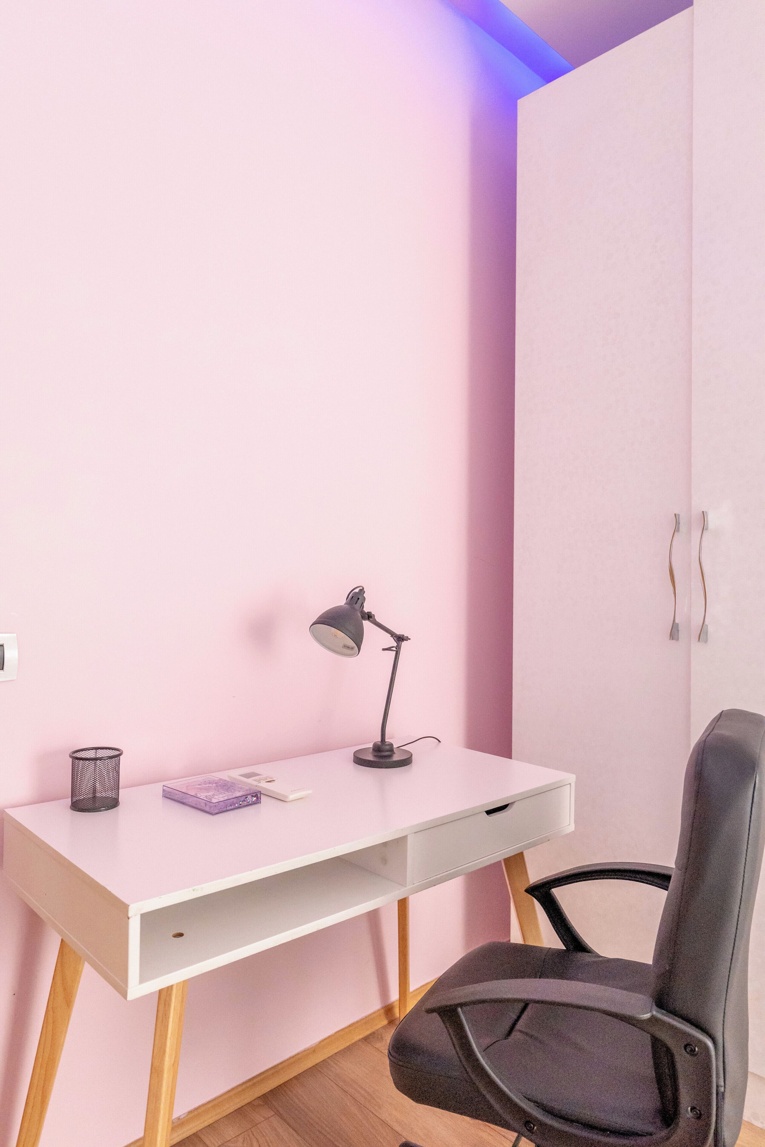Un escritorio blanco con una silla negra, una lámpara de escritorio y un libro morado.