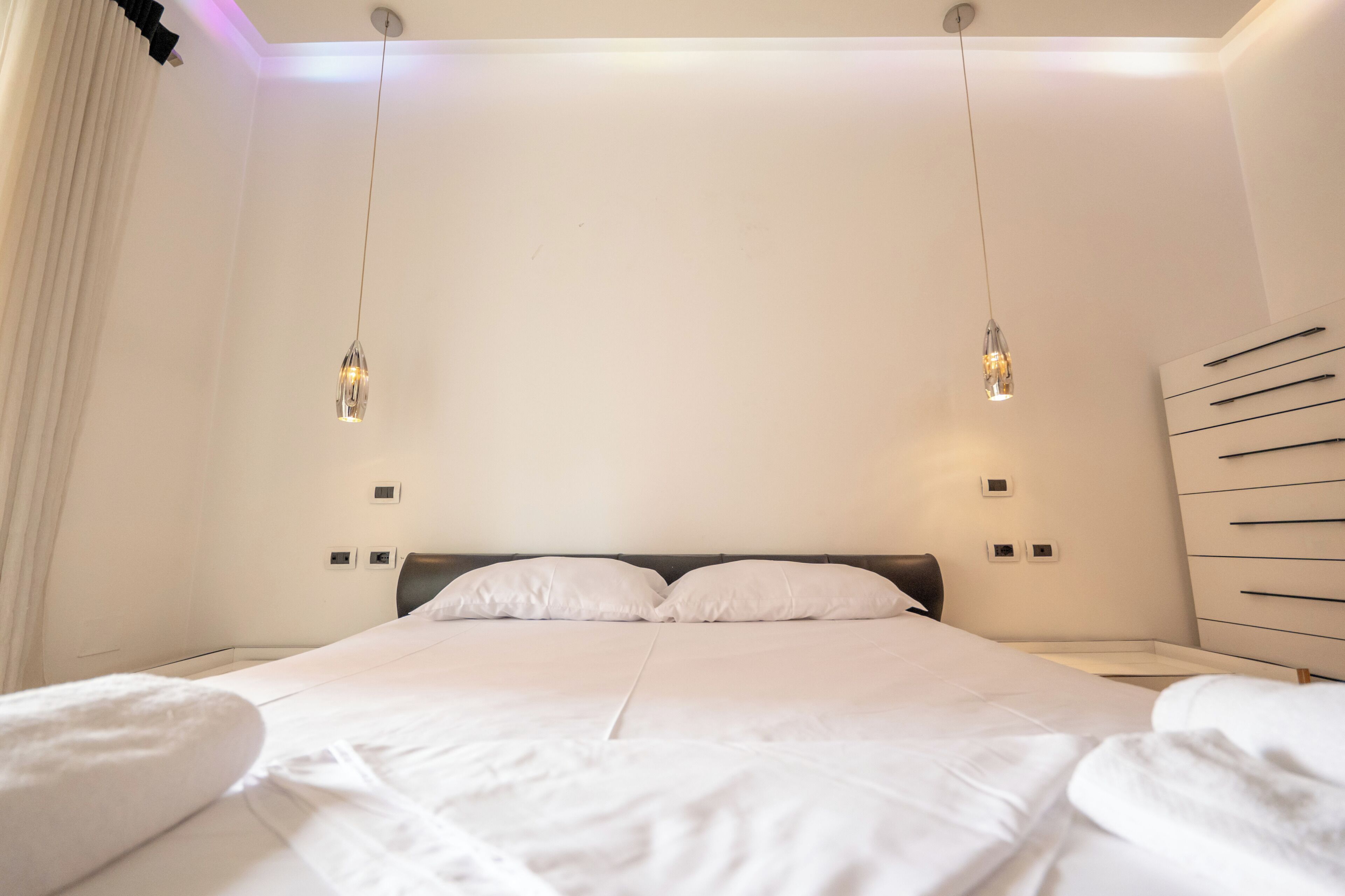Una habitación de hotel moderna con una cama grande, dos mesitas de noche, un aplique de pared y un banco empotrado con cajones.