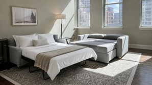 A bedroom with a bed, two sofas, a floor lamp, and large windows with city views.