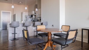 A modern dining area with a round wooden table and four chairs, a kitchen in the background, and pendant lighting.