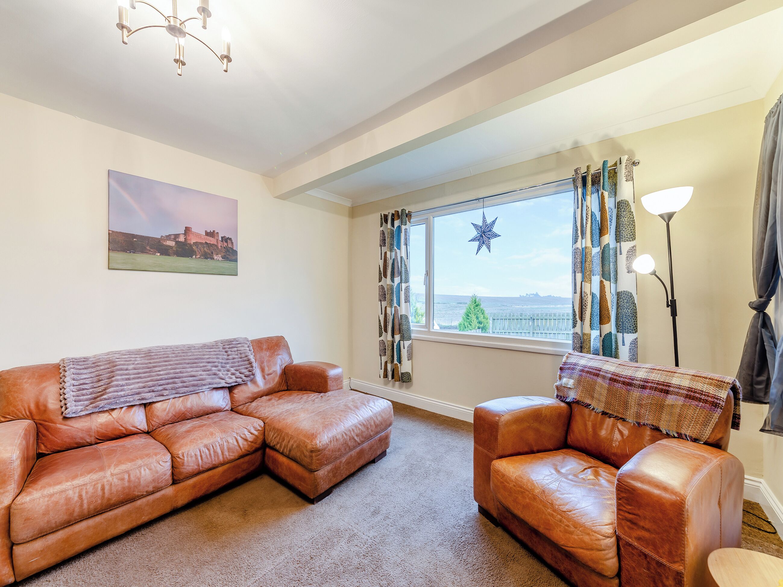 A living room with a brown leather sofa, a smaller armchair, a large window with curtains, a star decoration, and a painting on the wall.