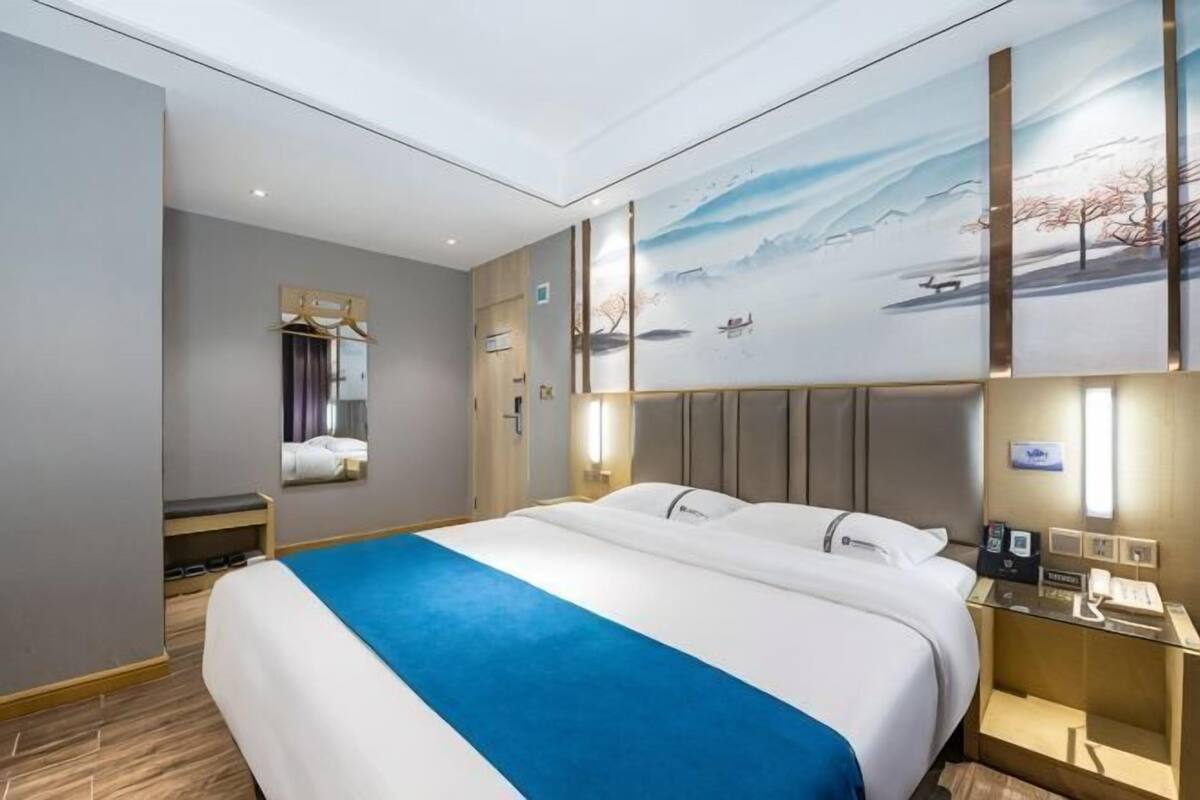 Habitación de hotel con una cama grande, una mesita de noche con lámpara, un espejo y una obra de arte en la pared que muestra un paisaje nevado.