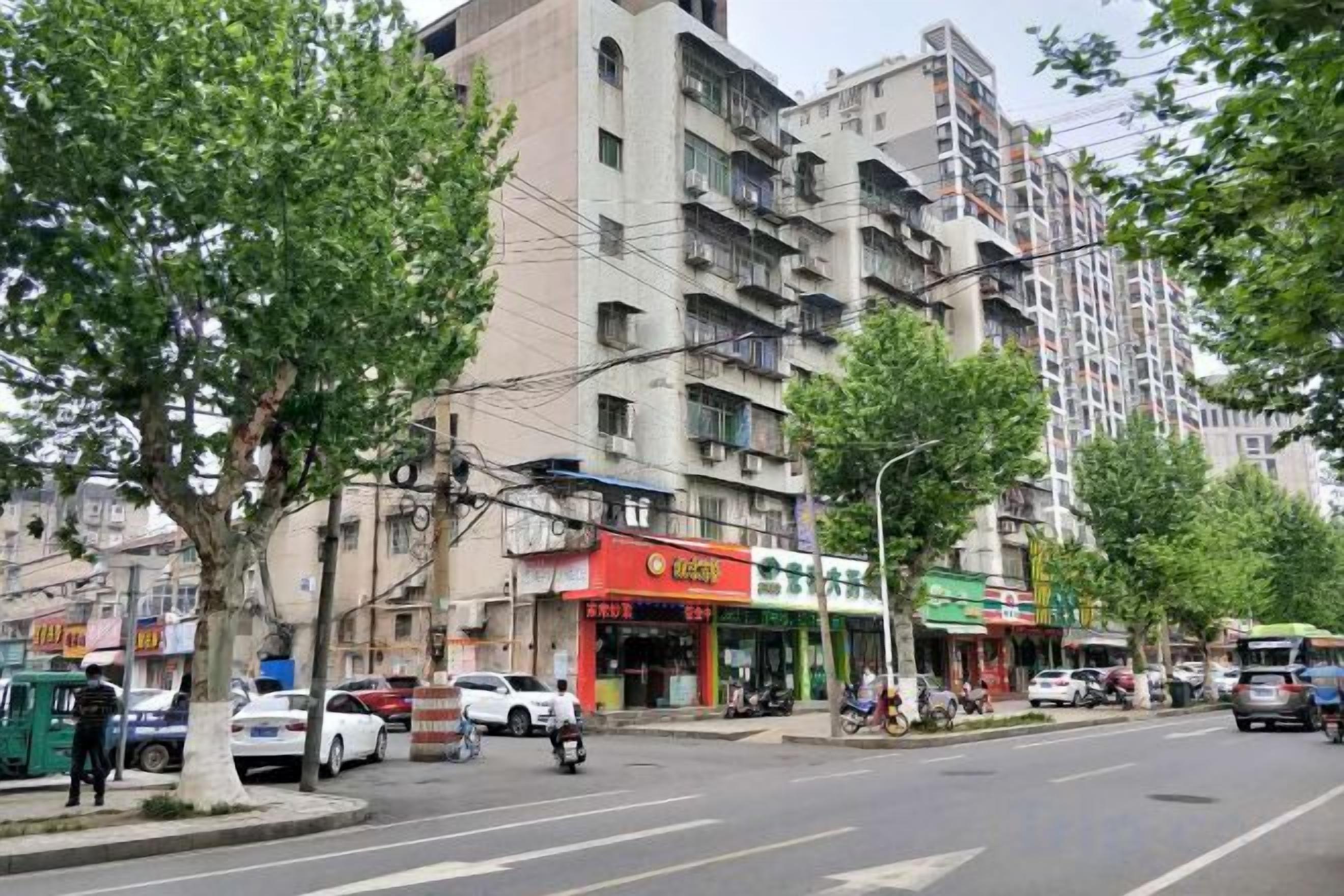 A street with multi-story buildings, parked cars, and trees.