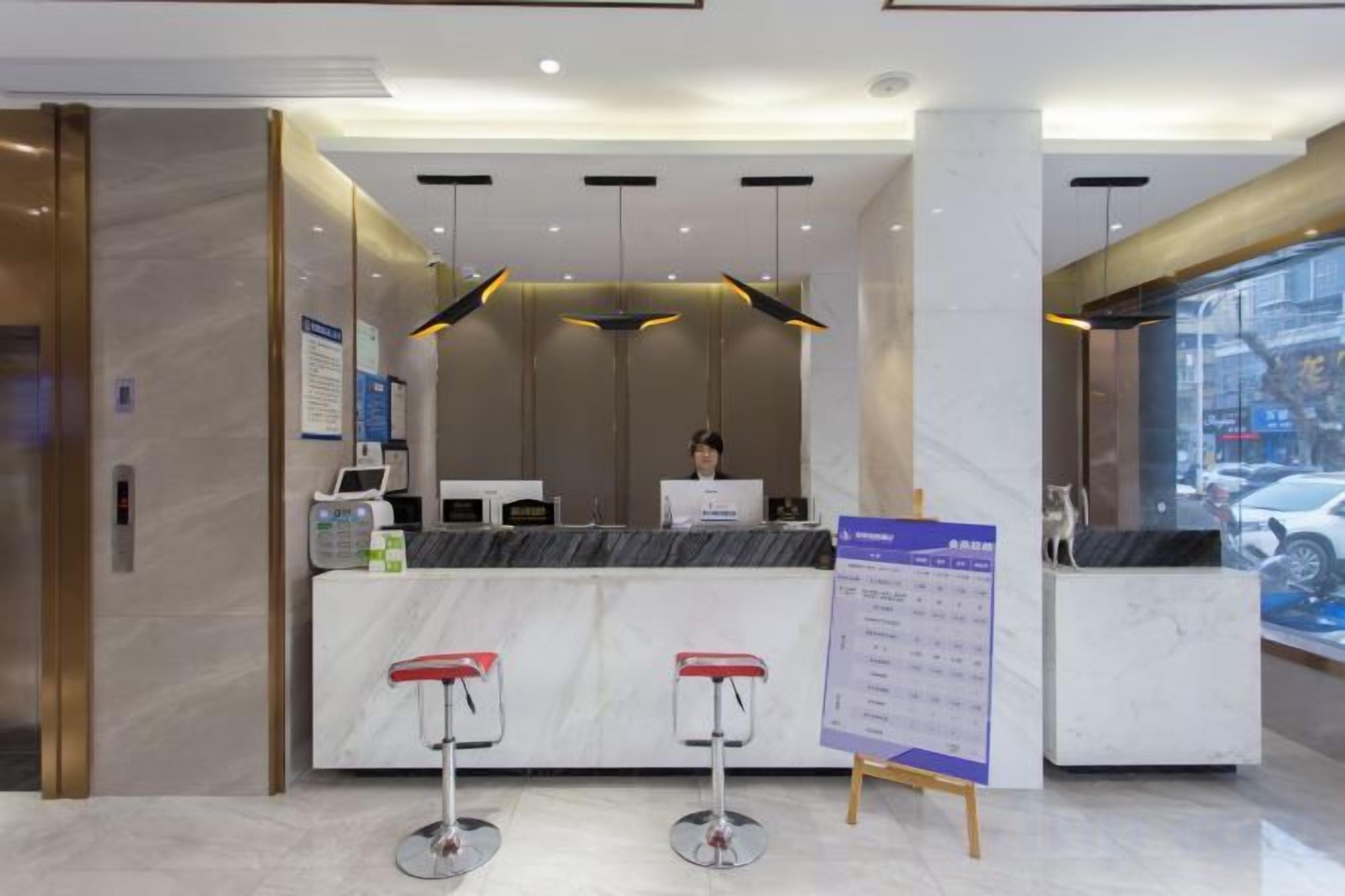 A hotel reception area with a marble counter, two red bar stools, a computer, and a signboard.