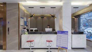 A hotel reception area with a marble counter, two red bar stools, a computer, and a signboard.