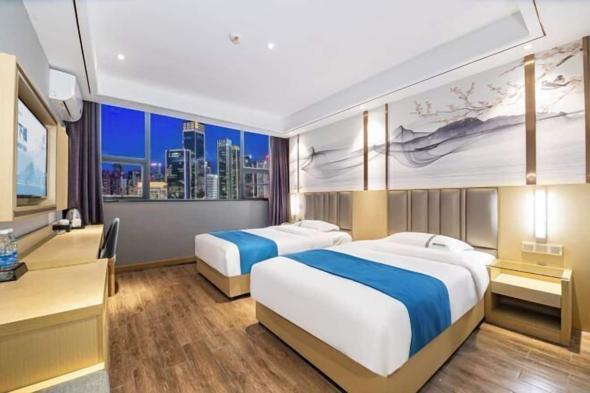 Habitación de hotel con dos camas, un escritorio, una silla, un ventanal con vista a la ciudad y un cuadro en la pared.