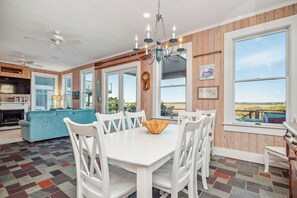 Amplia sala de estar con mesa de comedor, sillas blancas, lámpara de araña y vistas al exterior.
