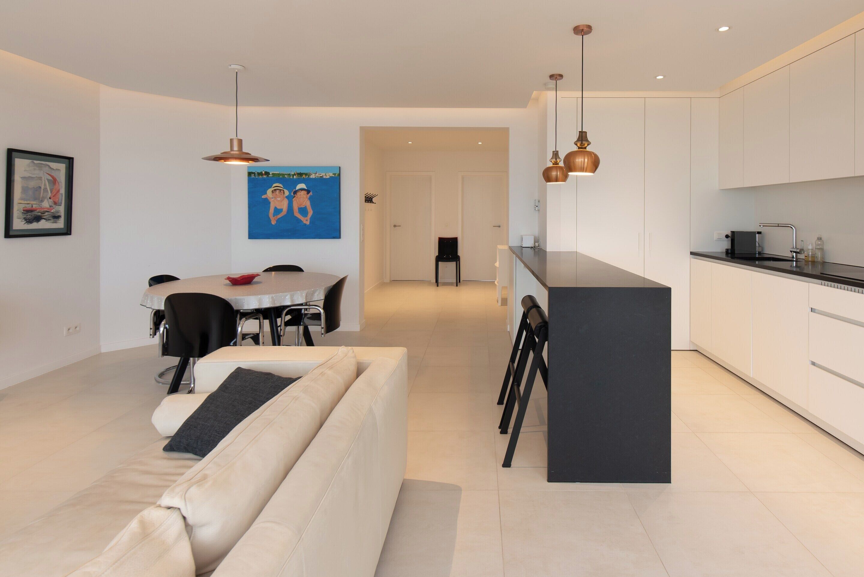 A modern kitchen with white cabinets and a black countertop, a dining area with a round table and chairs, and a beige sofa in the foreground.
