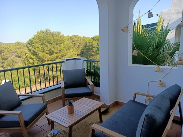 A balcony with a view of a forest, a table with a potted plant, and two chairs.