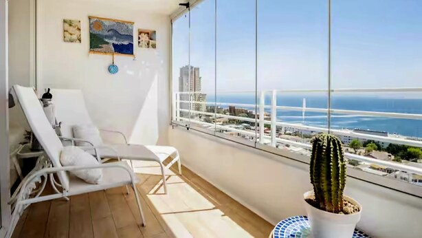 A balcony with a white metal chair, a potted cactus, and a view of the sea and cityscape.