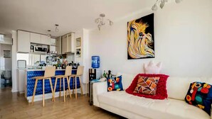 A modern living room with a white sofa, colorful cushions, and a kitchen area with a blue backsplash and white cabinets.