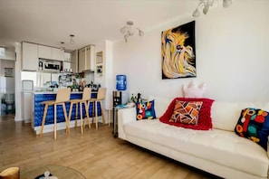 Un salon moderne avec un canapé blanc, des coussins colorés et un coin cuisine doté d’un plan de travail bleu et de meubles blancs.