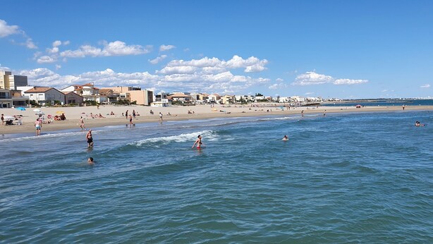 A beach with people swimming and sunbathing, buildings in the background, and a clear sky.
