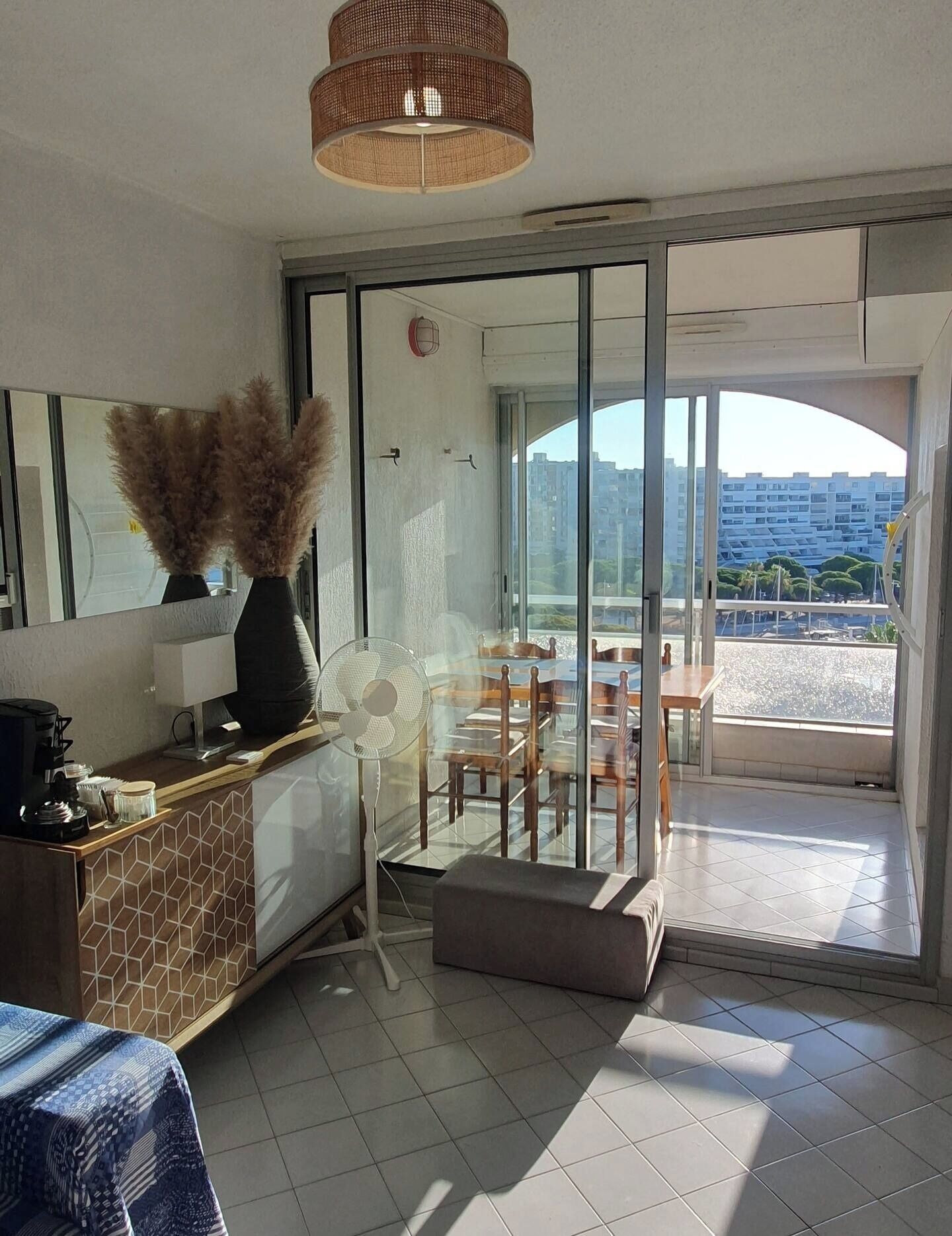 Ein modernes Hotelzimmer mit Glastür zu einem Balkon, einem Ventilator, einer Kaffeestation und Blick auf die Stadt.