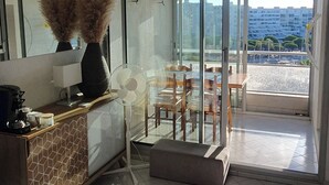 A modern hotel room with a glass door leading to a balcony, a fan, a coffee station, and a view of the cityscape.
