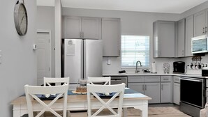 A modern kitchen with grey cabinets, a stainless steel refrigerator, and a dining area with white chairs and a wooden table. - Sea La (Port St. Joe)
