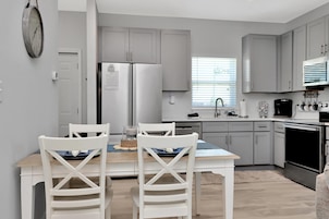 A modern kitchen with grey cabinets, a stainless steel refrigerator, and a dining area with white chairs and a wooden table.