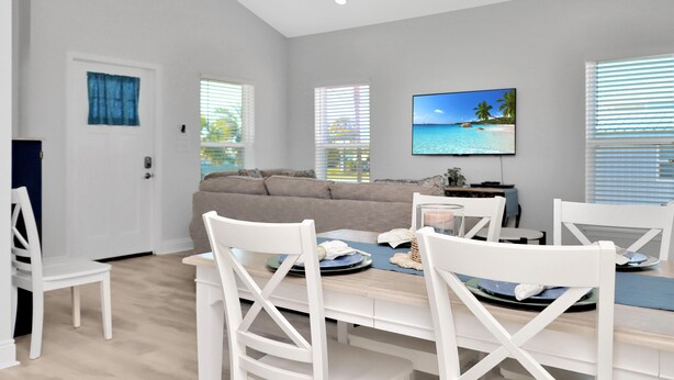A modern living room with a sofa, dining table, and chairs. There is a TV mounted on the wall displaying a beach scene. - Sea La (Port St. Joe)