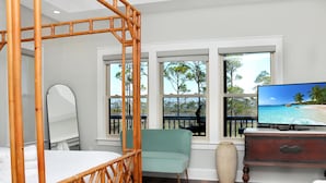 A bedroom with a four-poster bed, a television, a large window with a view of the beach, and a light blue chair. - Treetops (Port St. Joe)