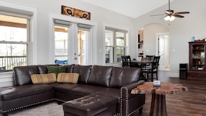 A spacious living room with a brown leather sectional sofa, a dining area with a wooden table, and a ceiling fan. - Treetops (Port St. Joe)