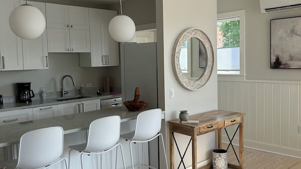 A modern kitchen with white cabinets, a central island with bar stools, pendant lighting, and a wooden dining table with a mirror above it. - Sunny downtown Belfast apartment with water view, right on Main St near eateries (Belfast)