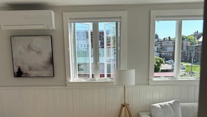 A living room with a wooden floor, a grey sofa, a grey armchair, and a floor lamp. There are two windows with a view of buildings and a car. - Sunny downtown Belfast apartment with water view, right on Main St near eateries (Belfast)