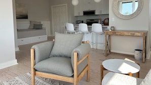 A modern, well-lit living room with a grey sofa, wooden coffee table, and a small round table. A kitchen area with white cabinets and a dining table with white chairs is visible in the background. - Sunny downtown Belfast apartment with water view, right on Main St near eateries (Belfast)