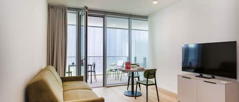 A modern living room with a sofa, a television on a cabinet, a dining area with chairs, and large windows.