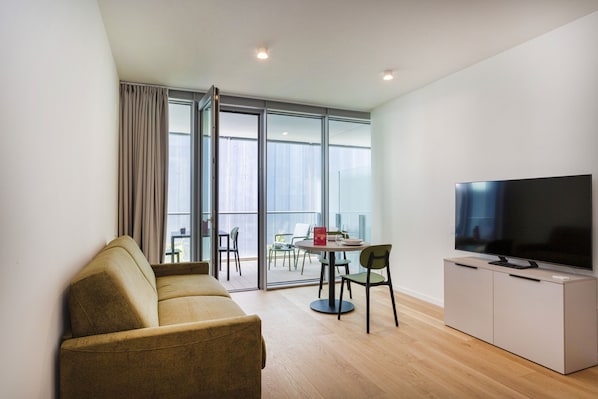 A modern living room with a sofa, a television on a cabinet, a dining area with chairs, and large windows.