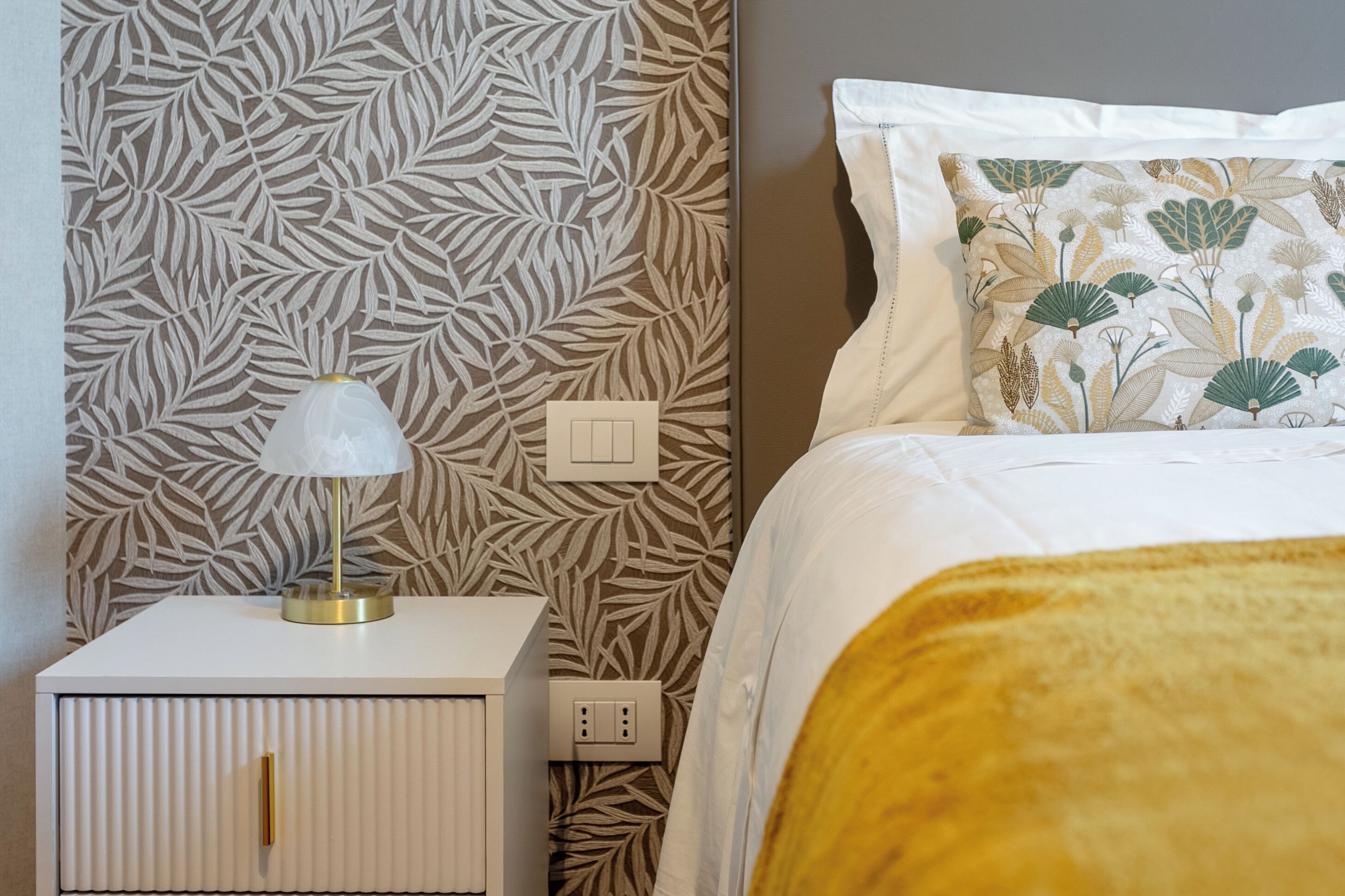 A neatly made bed with a white and yellow blanket, a white bedside table with a lamp, and a wall with a leaf pattern.