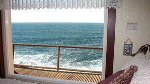A room with a view of the ocean through a window with a wooden deck. - Ocean Garden Suite at Whales Rendezvous (Depoe Bay)