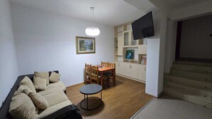 Un salon comprenant un canapé, une table à manger avec des chaises, une petite table ronde, un téléviseur fixé au mur et un escalier menant à un niveau supérieur.