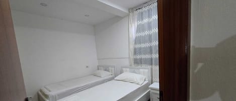 Una habitación pequeña y limpia con dos camas individuales, una mesita de noche y una ventana con cortinas.