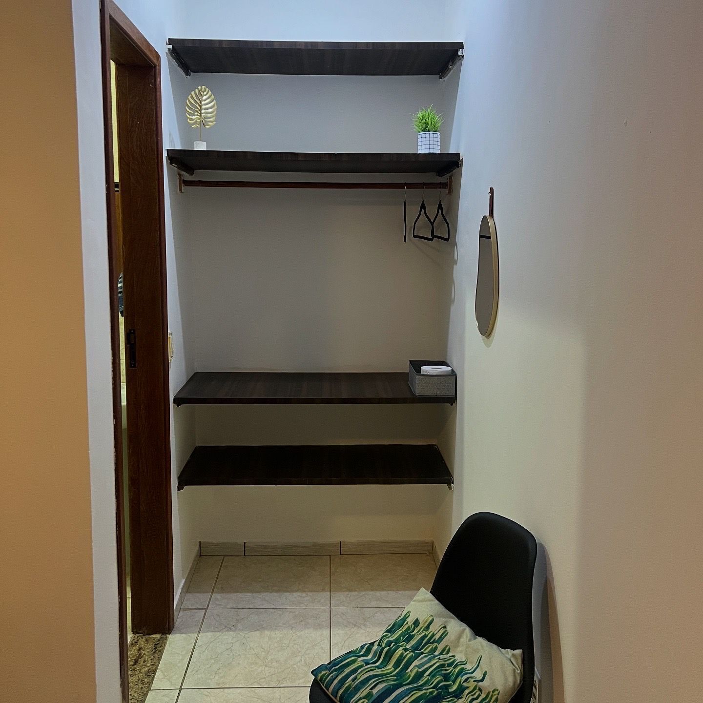 A small, narrow room with a built-in shelf unit against the wall, a chair with a patterned cushion, and a door leading to another area.