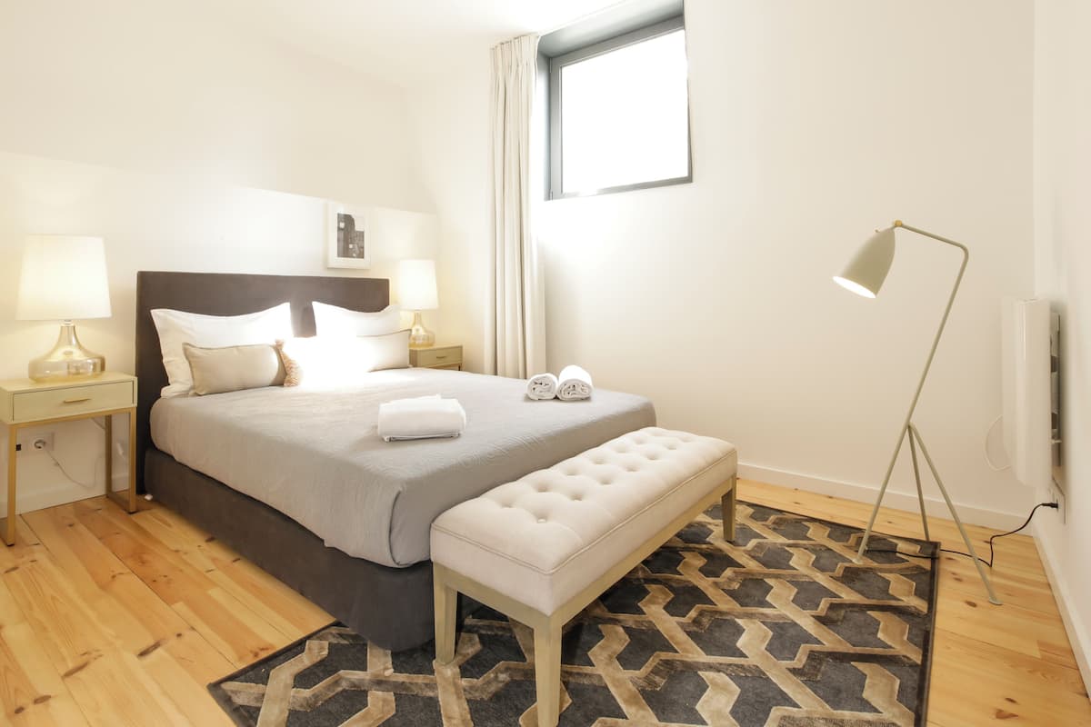 Une chambre à coucher bien rangée, avec un lit, des tables de chevet, un banc, une lampe sur pied et une fenêtre.