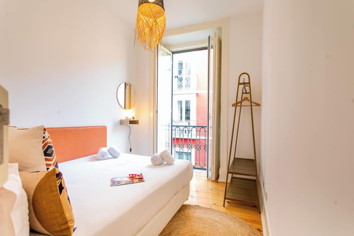 Une chambre d’hôtel comprenant un lit, une table de chevet, un miroir, un luminaire suspendu et une vue sur un balcon doté d’une balustrade.