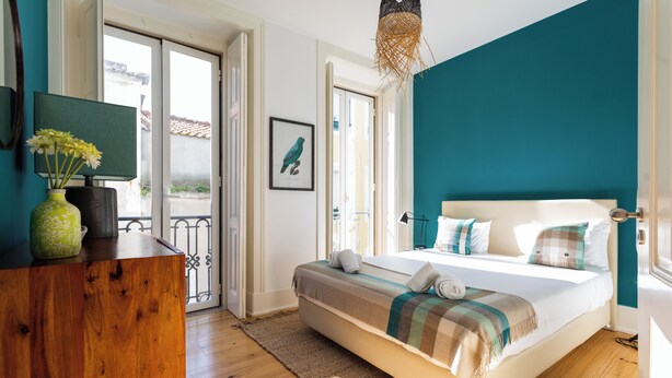 A bedroom with a bed, a wooden dresser, a vase with flowers, and a window with a view.