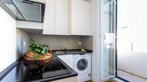 A compact kitchen with a washing machine, a basket of vegetables, and a sliding glass door leading to a balcony.
