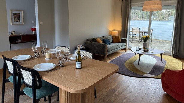 Ein modernes Wohnzimmer mit Essbereich, mit einem Holztisch, der für eine Mahlzeit gedeckt ist, einer Couch und einem kleinen runden Tisch mit einer Lampe.