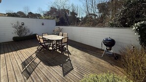 Eine Holzterrasse mit einem runden Tisch und Stühlen, einem Grill und einer weißen Wand.